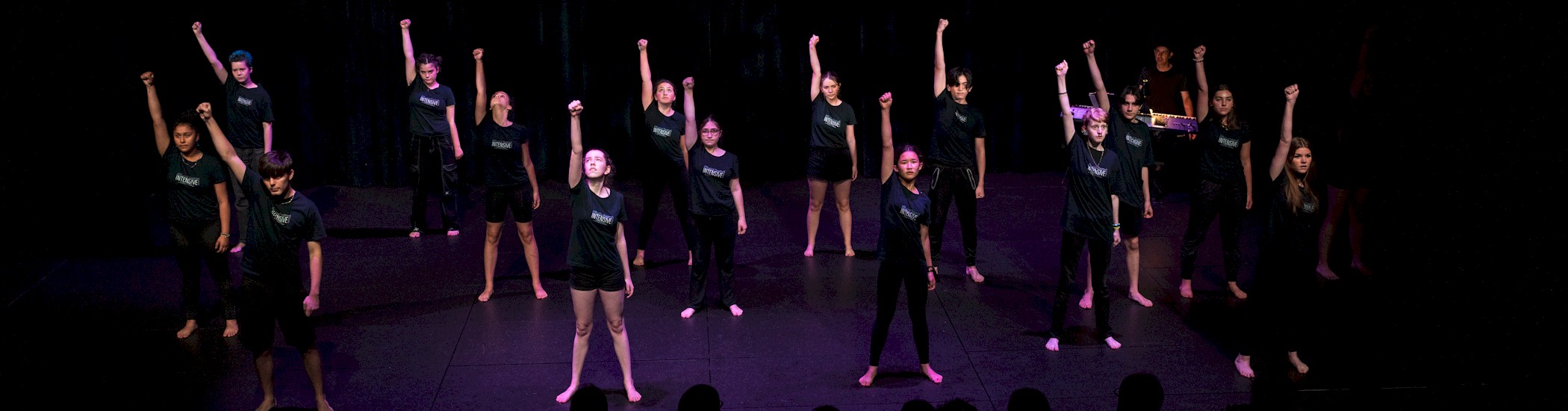 Photo of young artists in black clothing performing on a dimly lit stage.