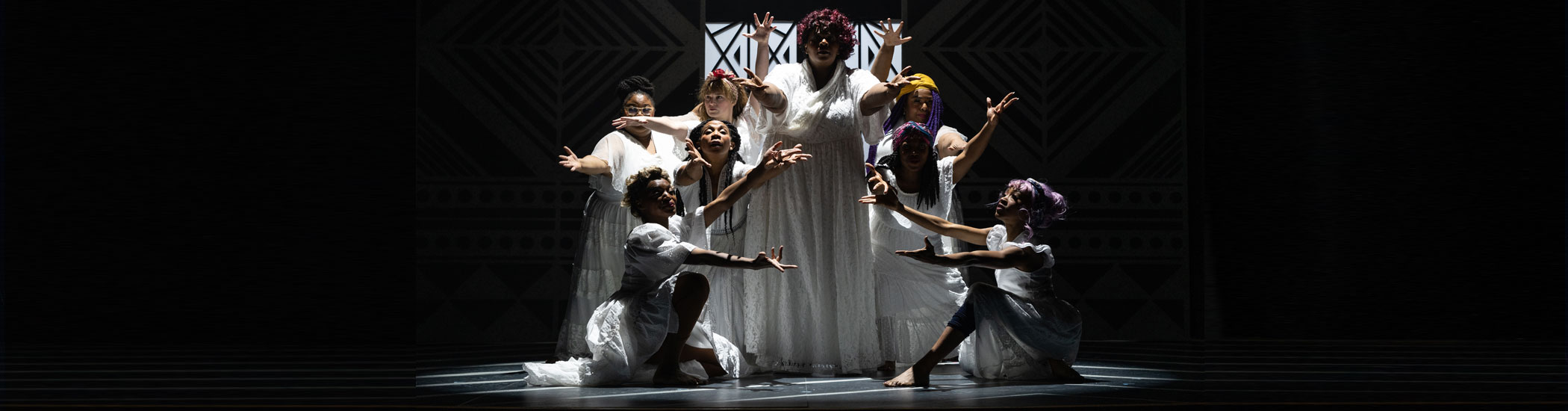 Eight women in long white dresses pose in a semicircle on a darkened stage