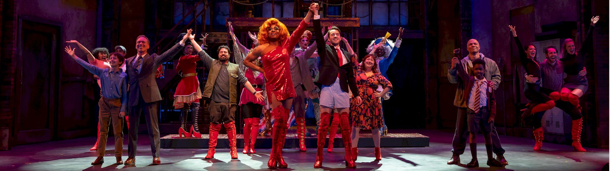 Cast members of the musical Kinky Boots onstage with their arms raised in joy