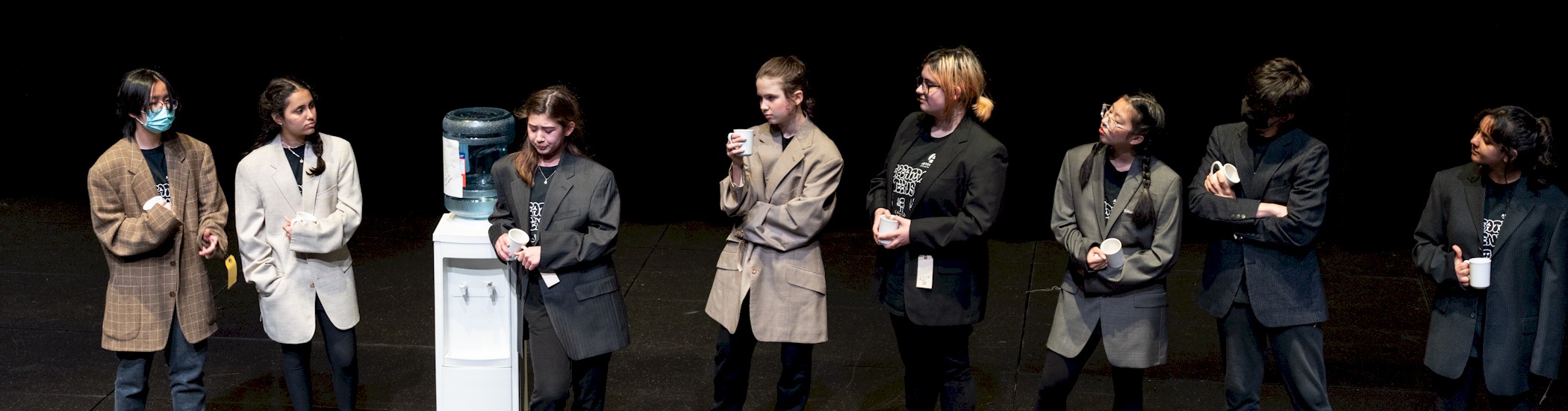 A group of nine teens perform onstage wearing business suits and standing in a row next to a water cooler