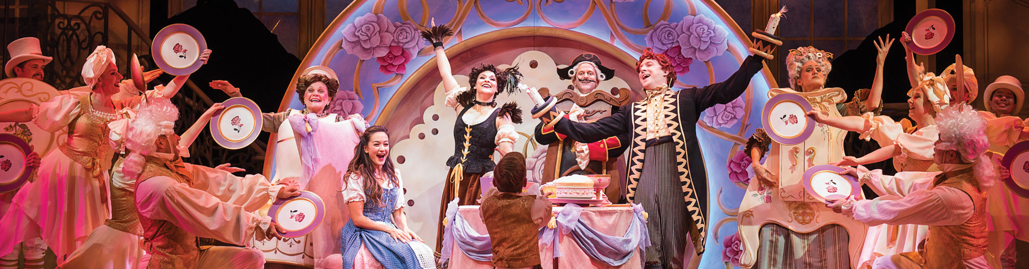 Photo of a smiling Belle sitting onstage surrounded by Lumière, Cogsworth, Mrs. Potts, Featherduster, and Wardrobe. 