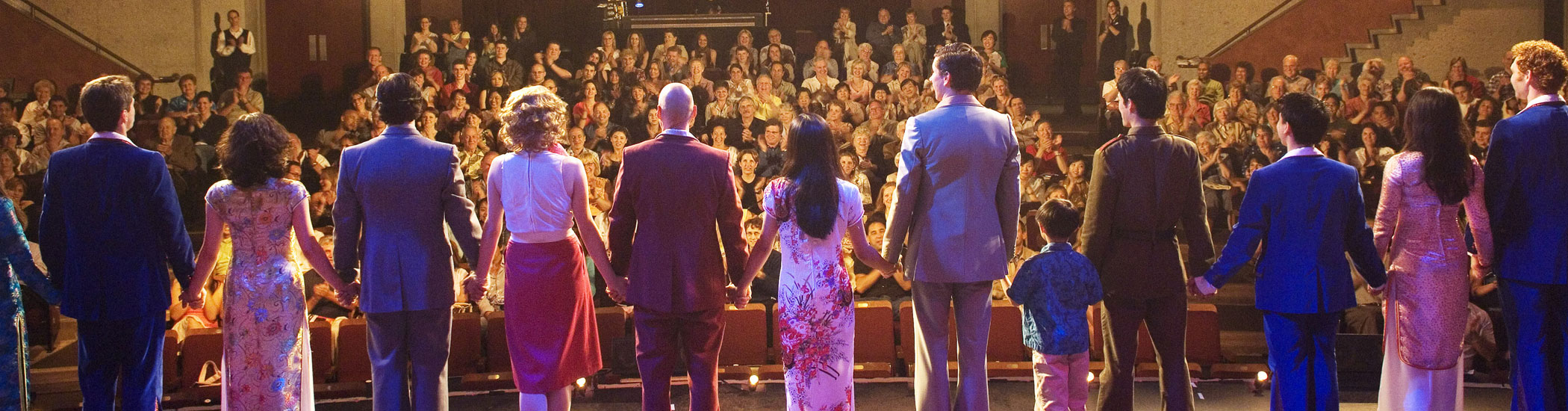 Photo from behind of a cast taking their final bow onstage, highlighting the applauding audience.