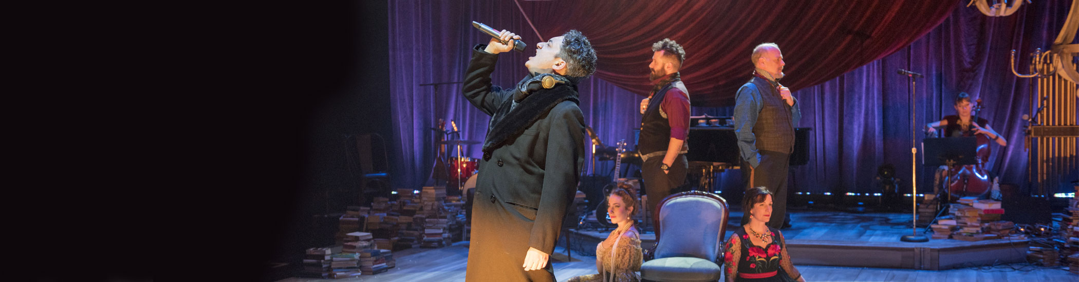Photo of an actor onstage is singing into a mic with four actors behind him and a cellist in the background.