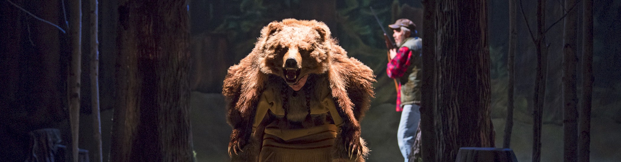 Photo of an actor onstage with a bear suit over their back. Another actor in hunting gear steps out from behind a tree.