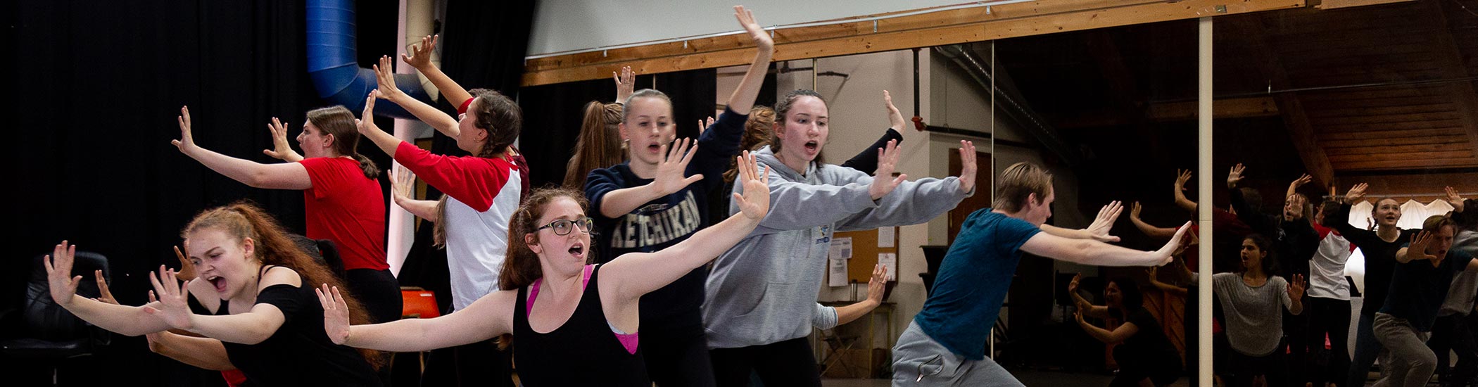 Photo of students rehearsing in a studio. They are in mid-movement, with their mouths open singing.
