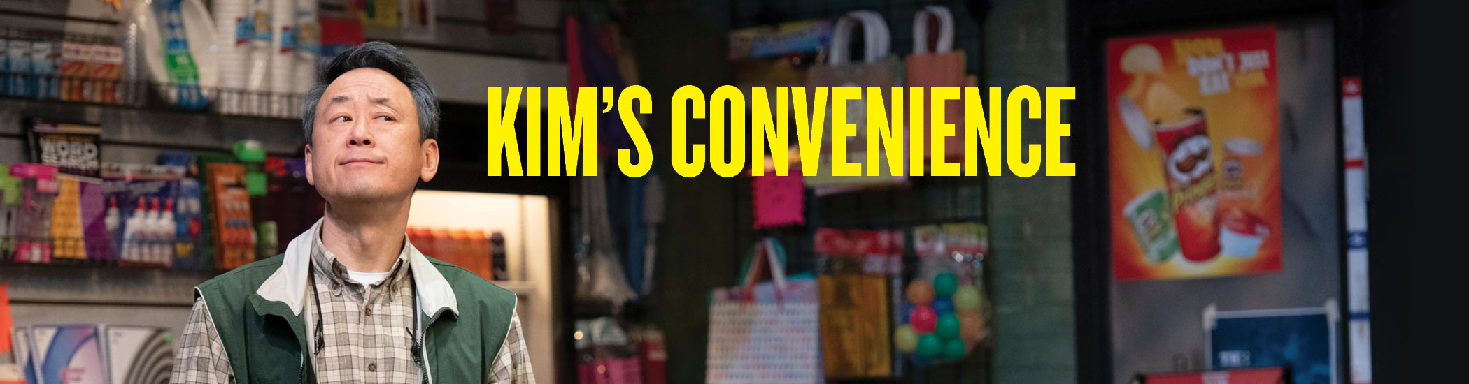 Yellow Text reads “Kim’s Convenience” overlayed on a photo of a Korean-Canadian shopkeeper. He is wearing a green vest and brown button up, standing in a convenience store and looking out of the corner of his eye at something out of frame.